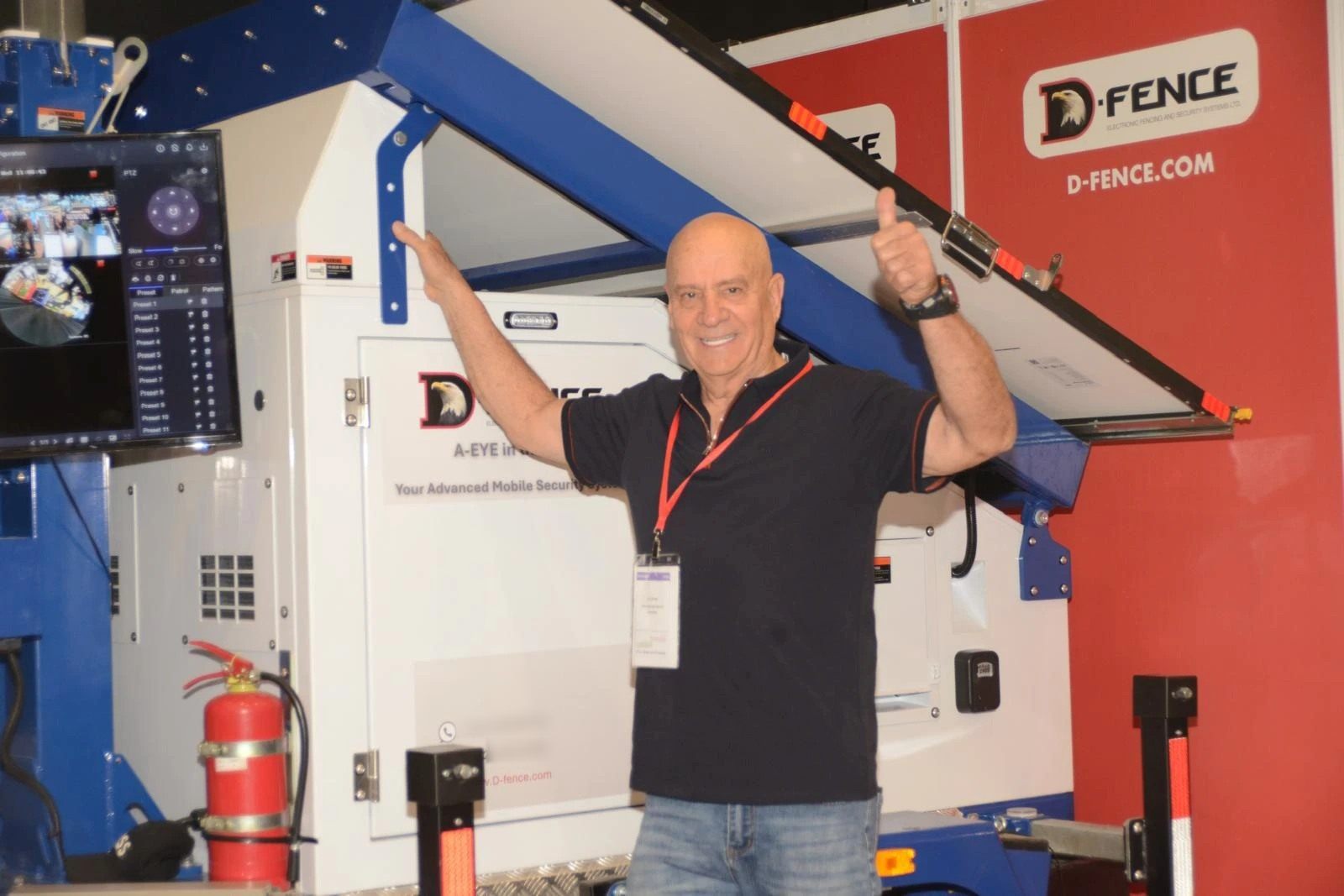 A man gives a thumbs-up beside the AI-EYE IN THE SKY solar trailer on the D-FENCE exhibition stand, next to the live camera control screen