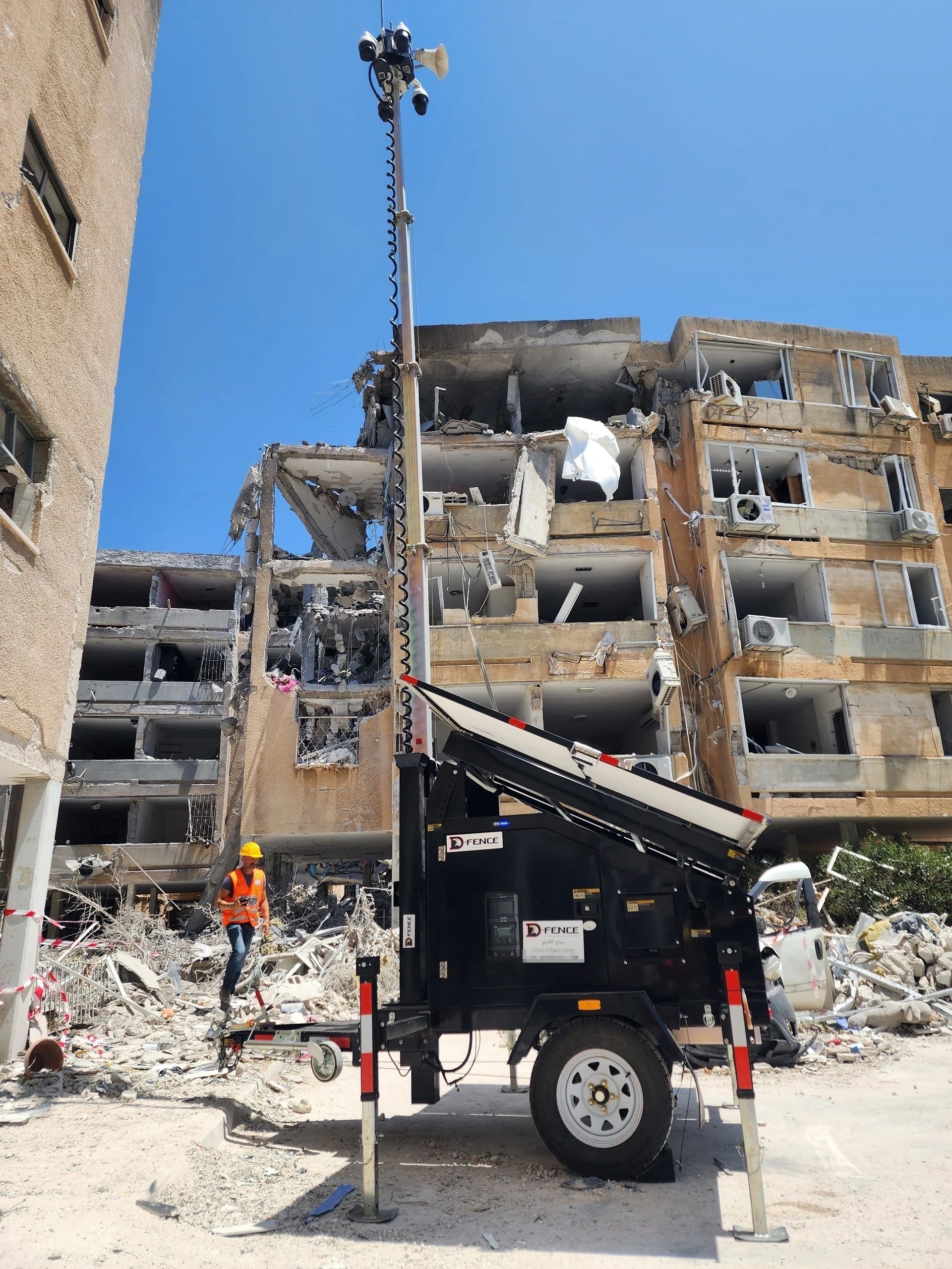 Black AI-EYE IN THE SKY trailer with its mast, PTZ cameras and loudspeaker raised beside a heavily damaged apartment block