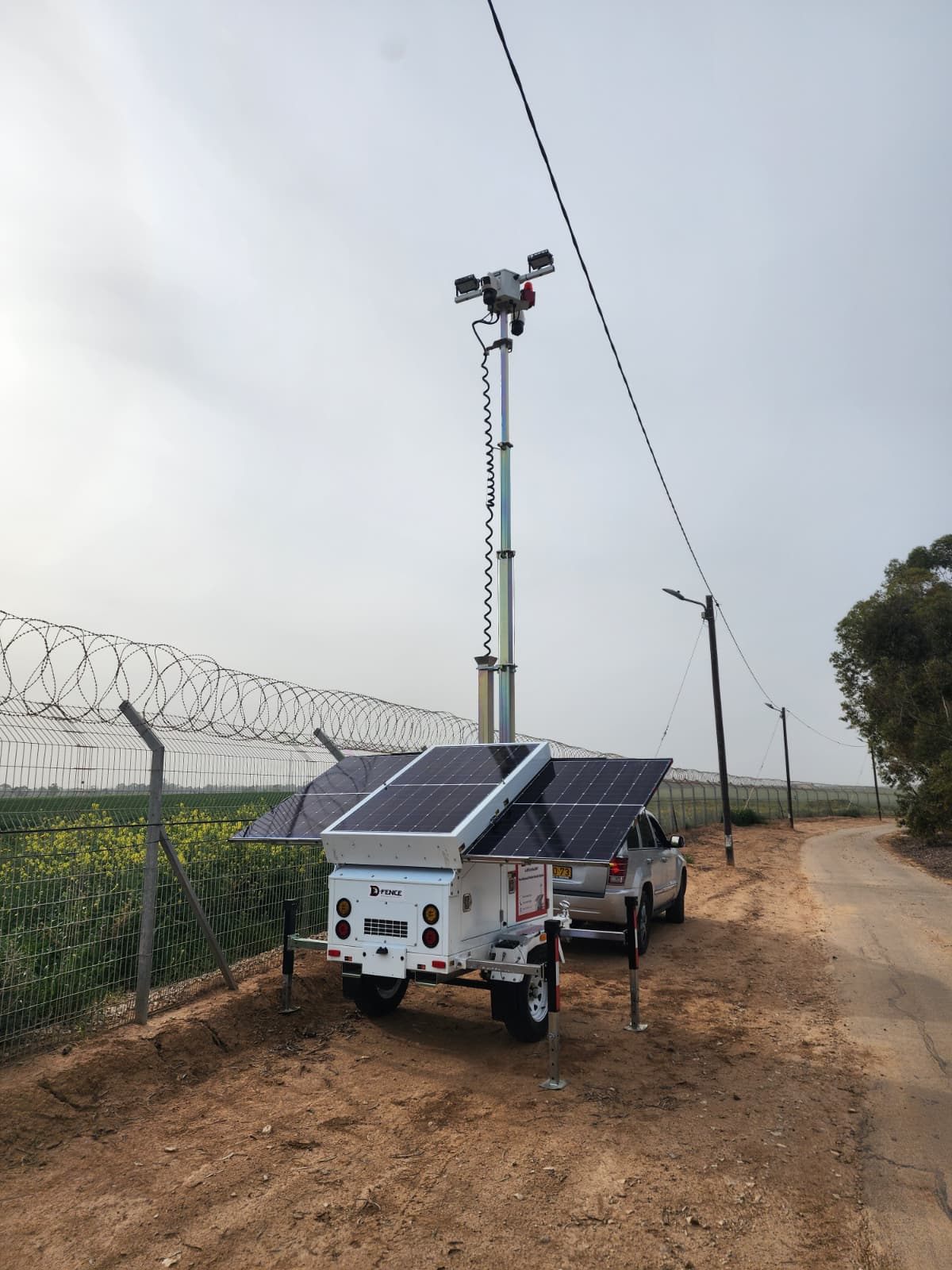 White AI-EYE trailer with floodlights and a PTZ camera on its raised mast, beside a concertina-topped fence and a patrol road
