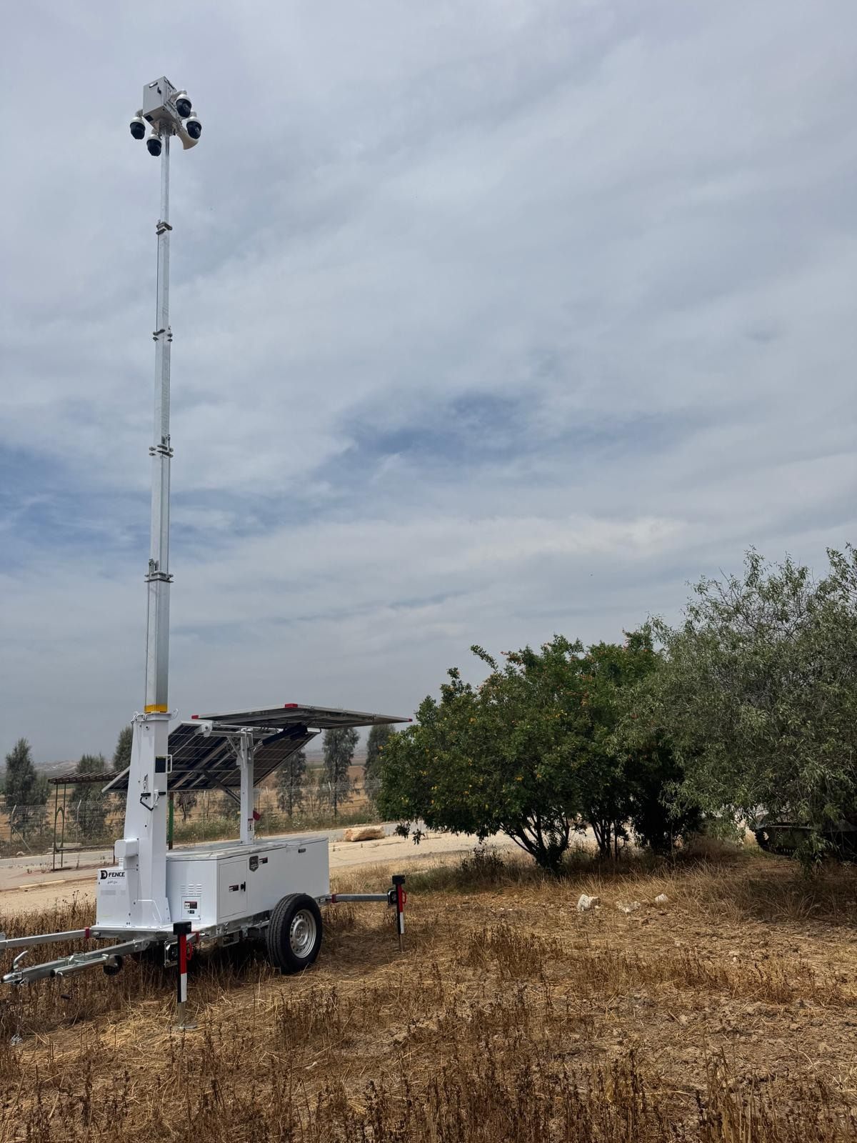 White AI-EYE trailer with a four-camera head on a raised mast in a dry field next to olive trees