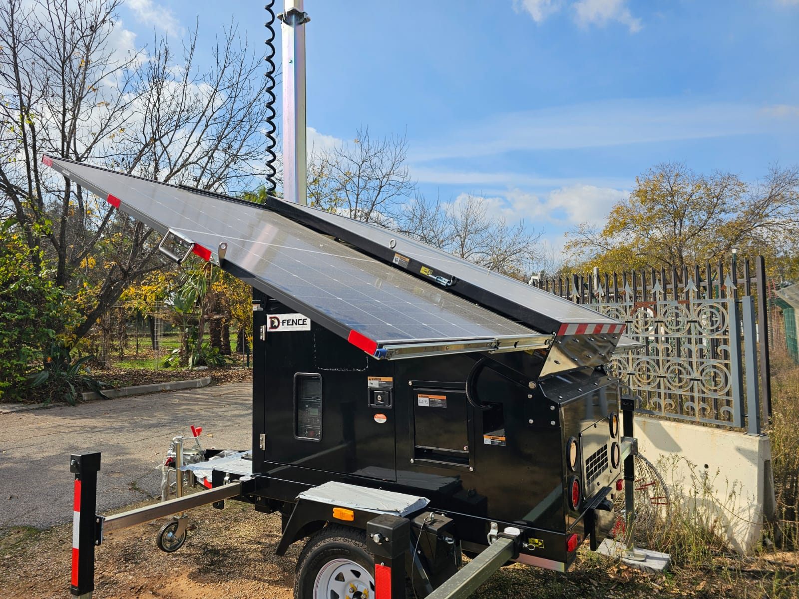 Black D-FENCE AI-EYE IN THE SKY solar trailer with its solar wings open and the telescopic mast rising into a blue sky