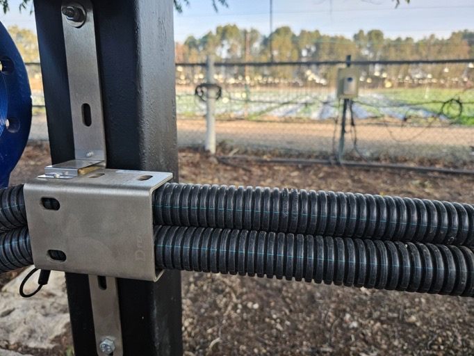 Steel D-FENCE clamp gripping two corrugated cable conduits on a post at an outdoor field installation