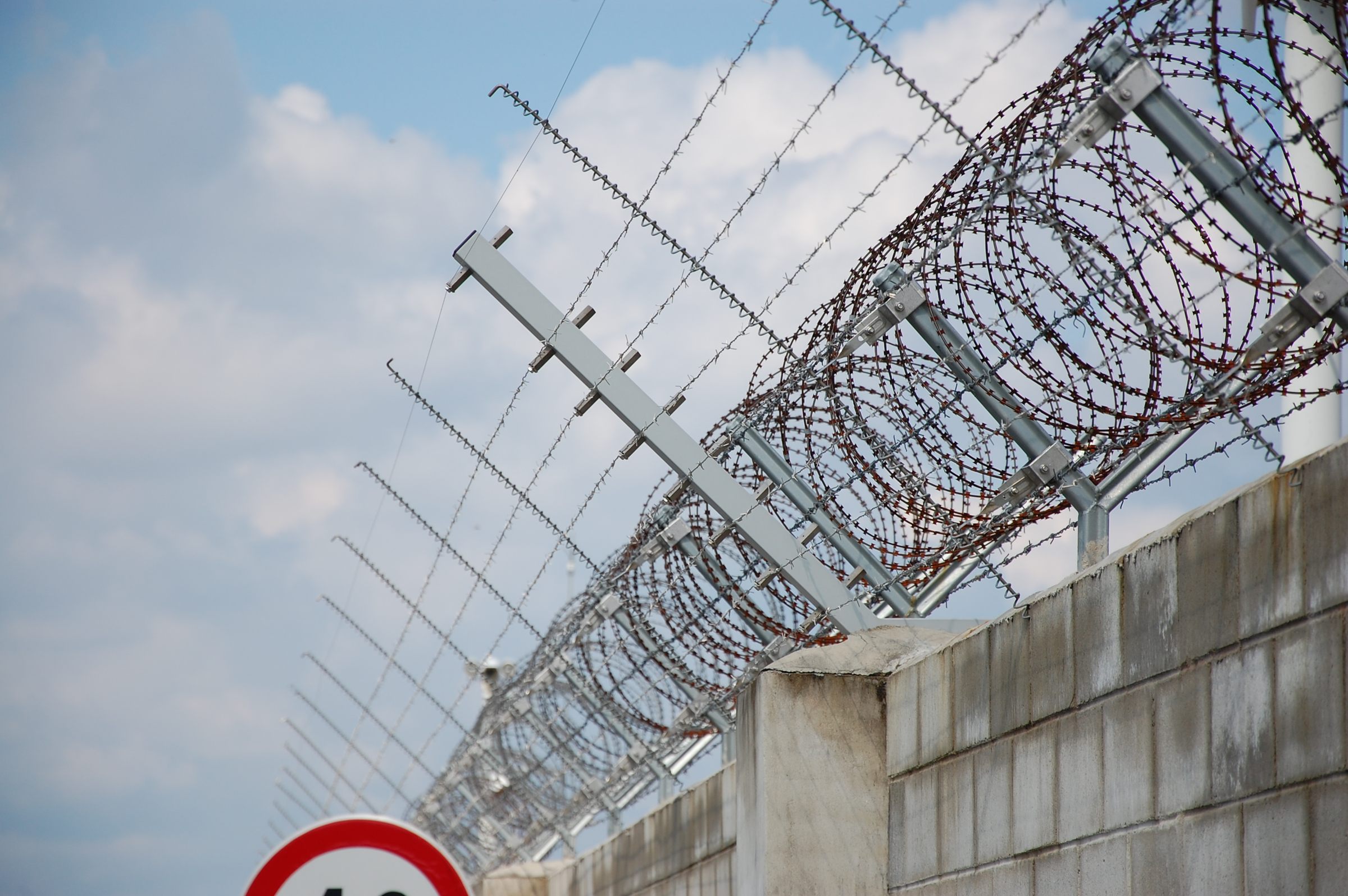 Razor-wire concertina on sensor arms and taut-wire outriggers along the crown of a block perimeter wall