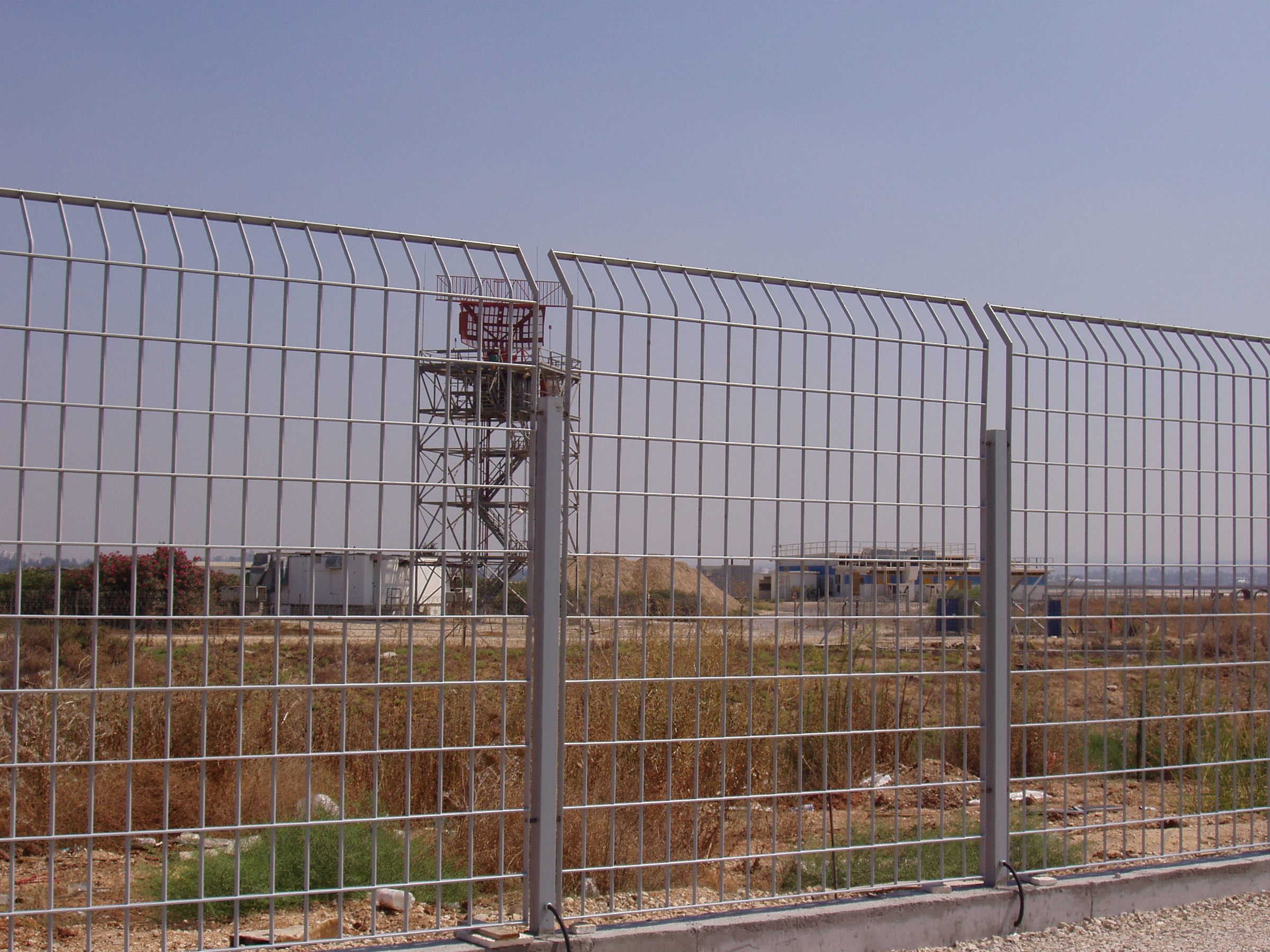 D-Corative panels and grey sensor posts, with sensor cabling at their base, on the airside perimeter of Ben Gurion Airport; a radar tower stands behind