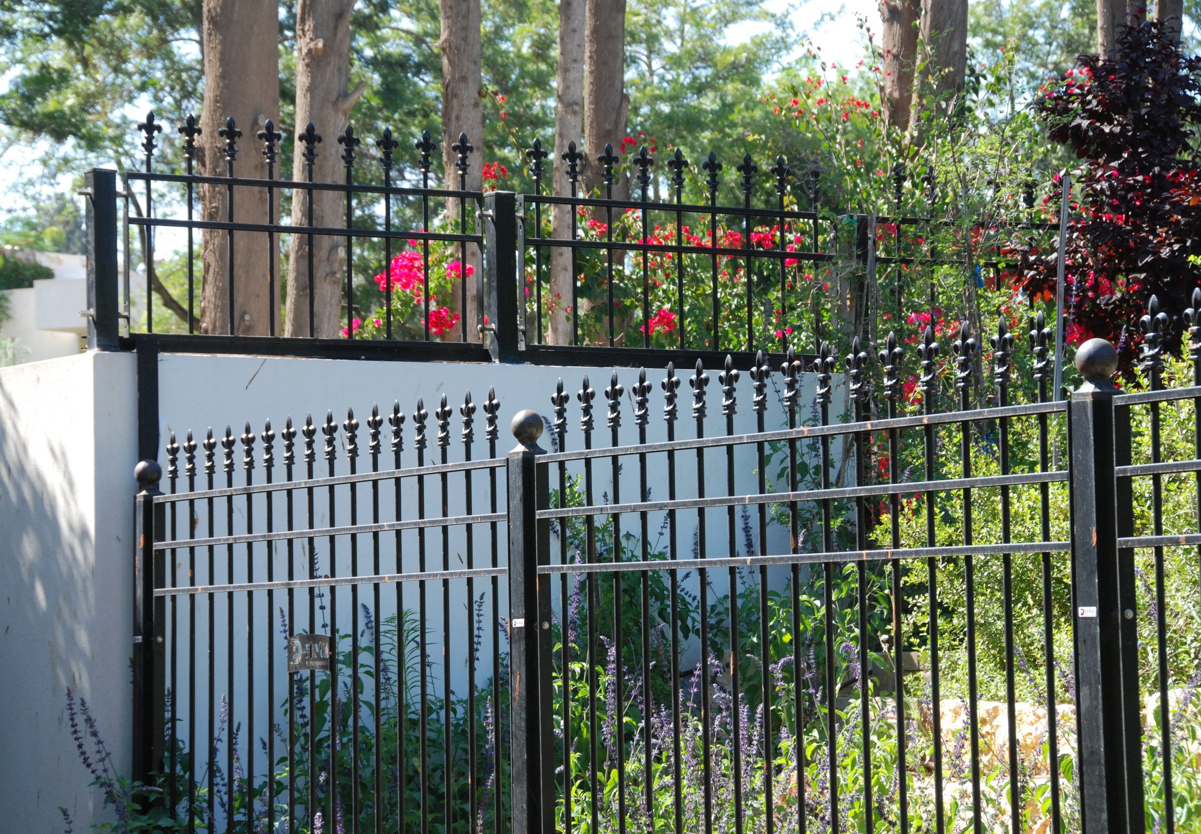 Fleur-de-lis ornamental fence on a white wall; stainless-steel sensor brackets join the post to the panel
