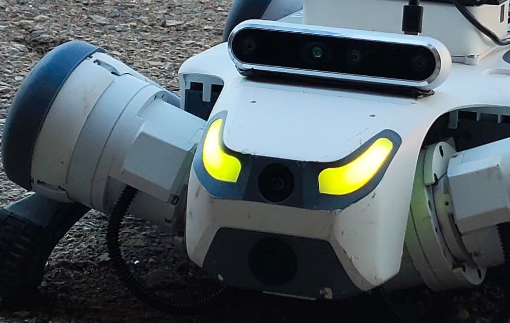 Close front view of D-DOG: depth-camera bar, front camera and lit status lights, with a compute module on its back