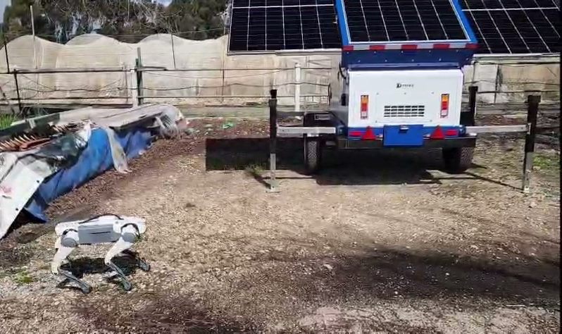 D-DOG walking in a yard in front of a D-FENCE solar surveillance trailer with open panels