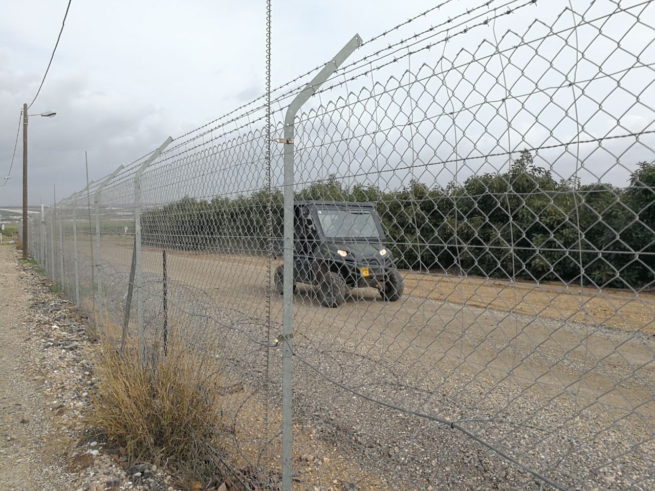 Field fence with a barbed-wire top along an agricultural perimeter road; a patrol vehicle drives inside the fence past an orchard