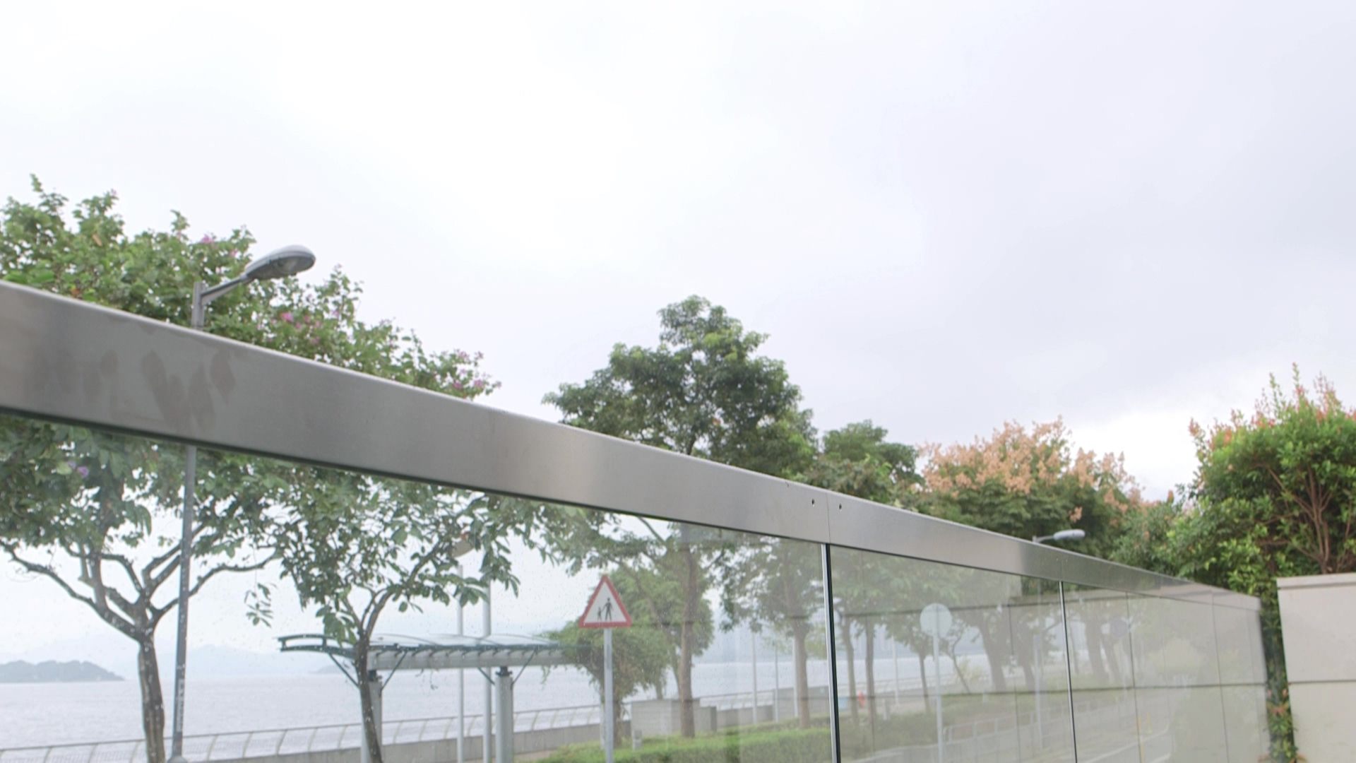Glass fence with a brushed-steel capping along a seafront road, trees and the sea behind