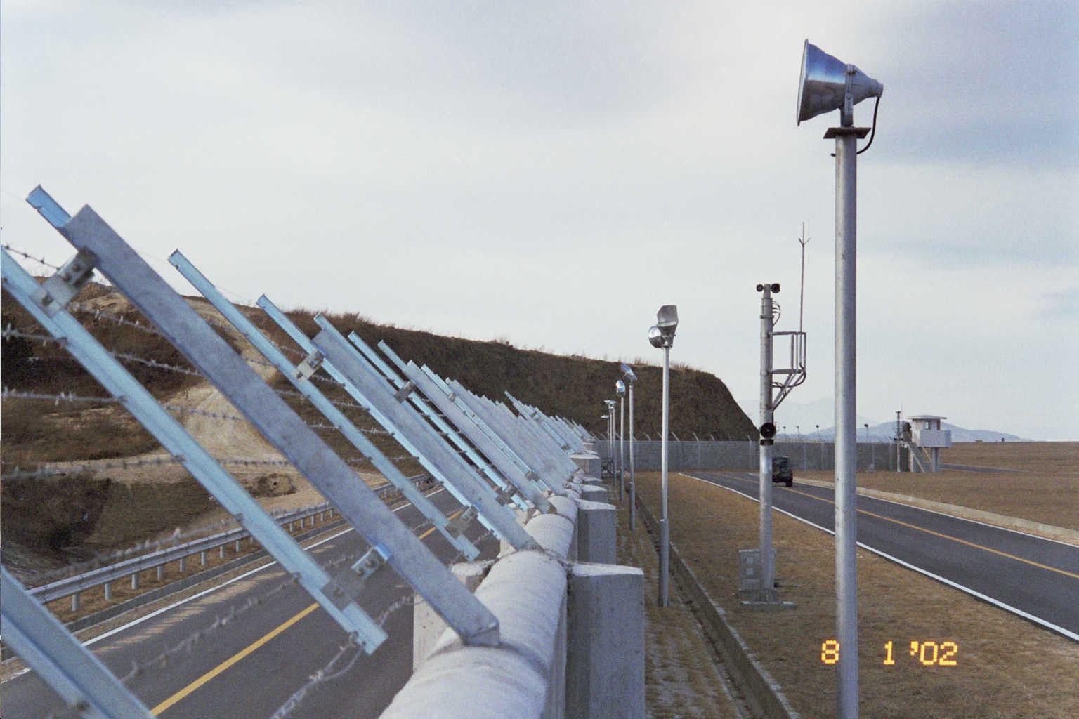 Angled outriggers carrying tensioned wires along the perimeter wall of Yang Yang International Airport, patrol road and lighting poles beside it
