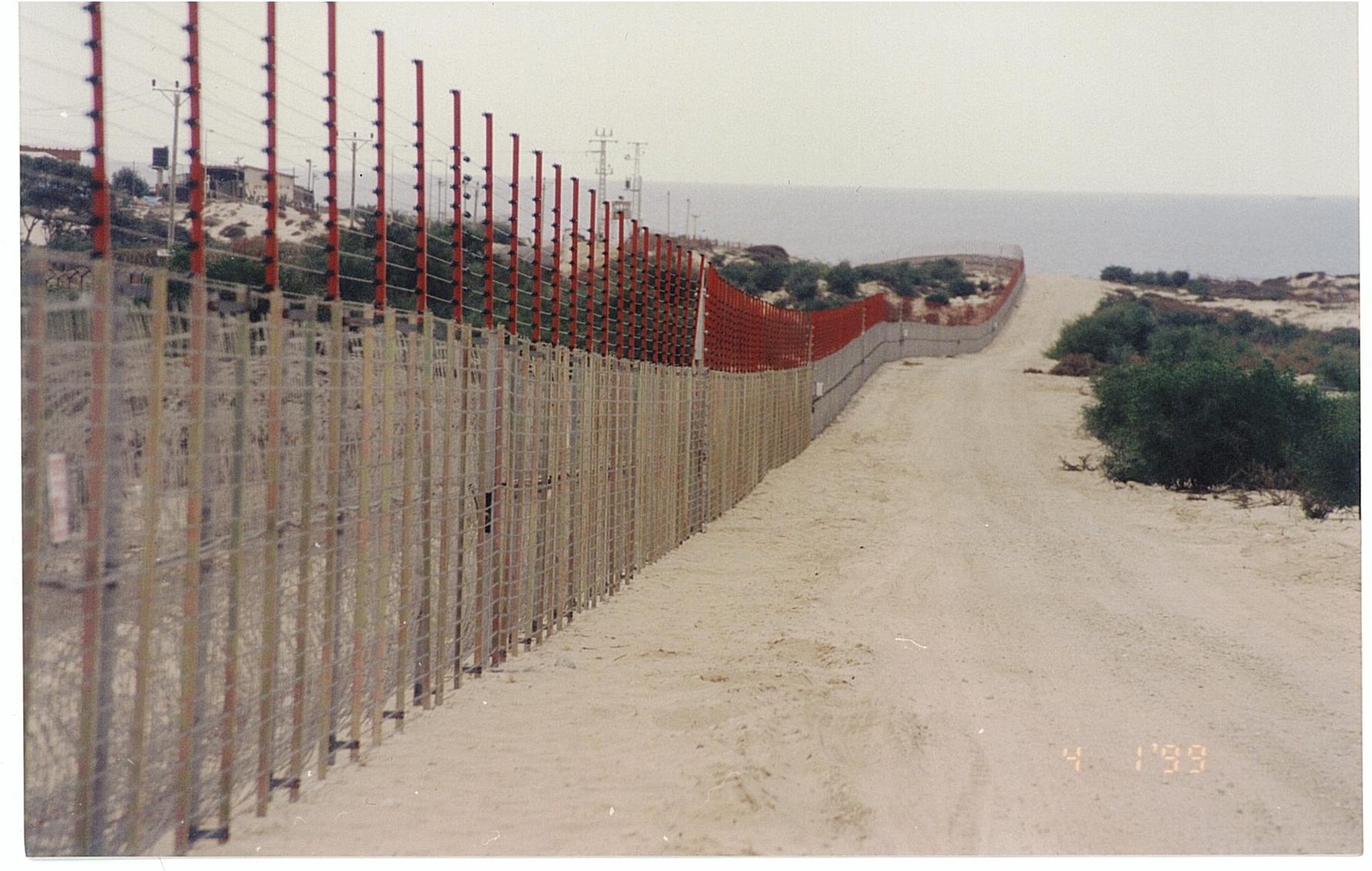 Archive photo, 1999: long taut-wire border fence with red-topped posts beside a sandy patrol road by the sea