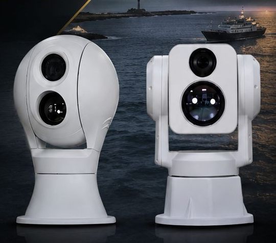 Two white maritime gyro-stabilised day/thermal camera turrets against a night sea with a lighthouse and a ship